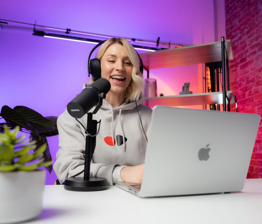 Podcast creator wearing headphones and speaking into a microphone beside a laptop